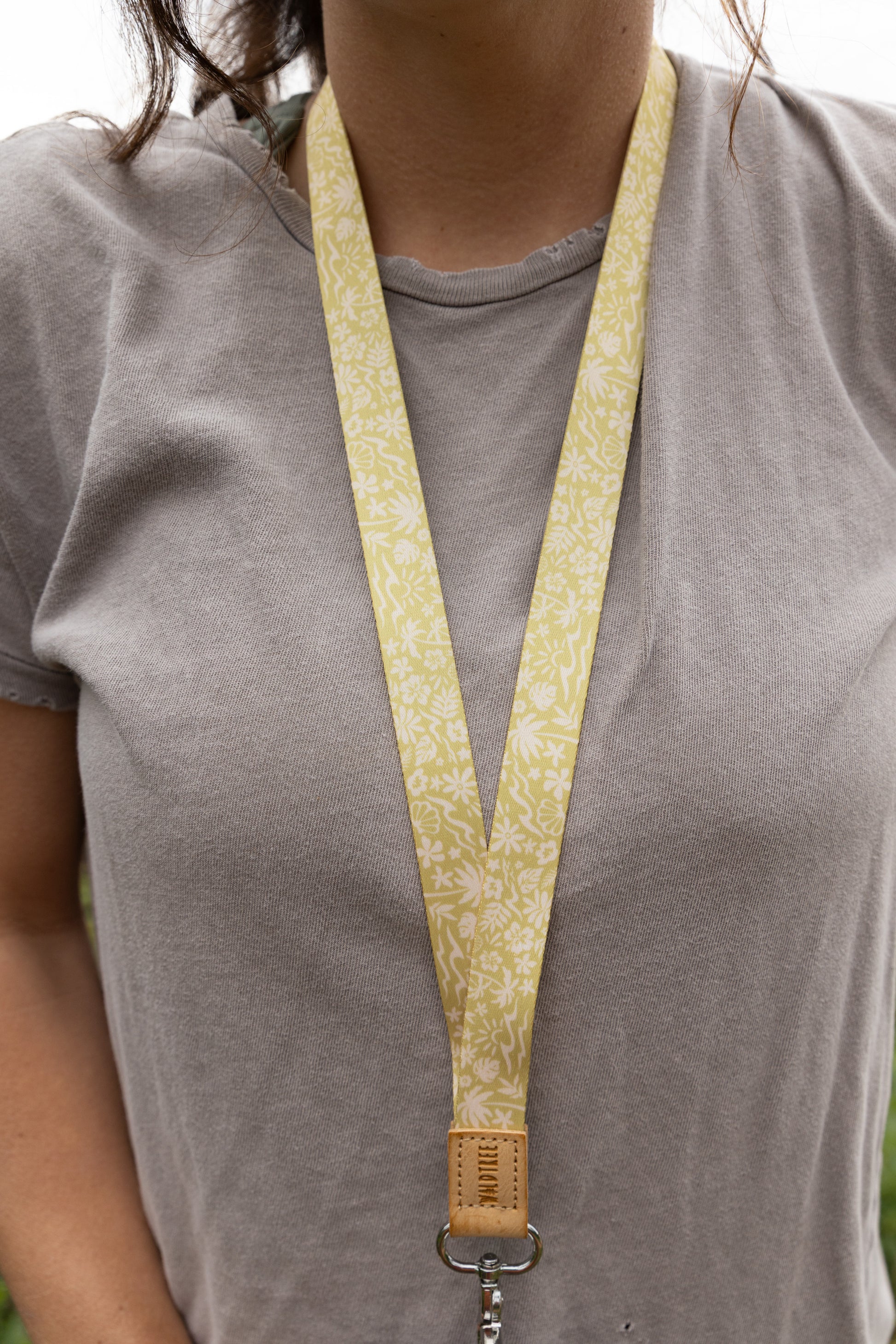 Close-up of the Sunny Day neck lanyard worn around a woman's neck, the yellow strap printed with waves, palm trees, and seashells forming a V shape against her gray t-shirt, ending in a tan leather WILDTREE tab and silver clasp.
