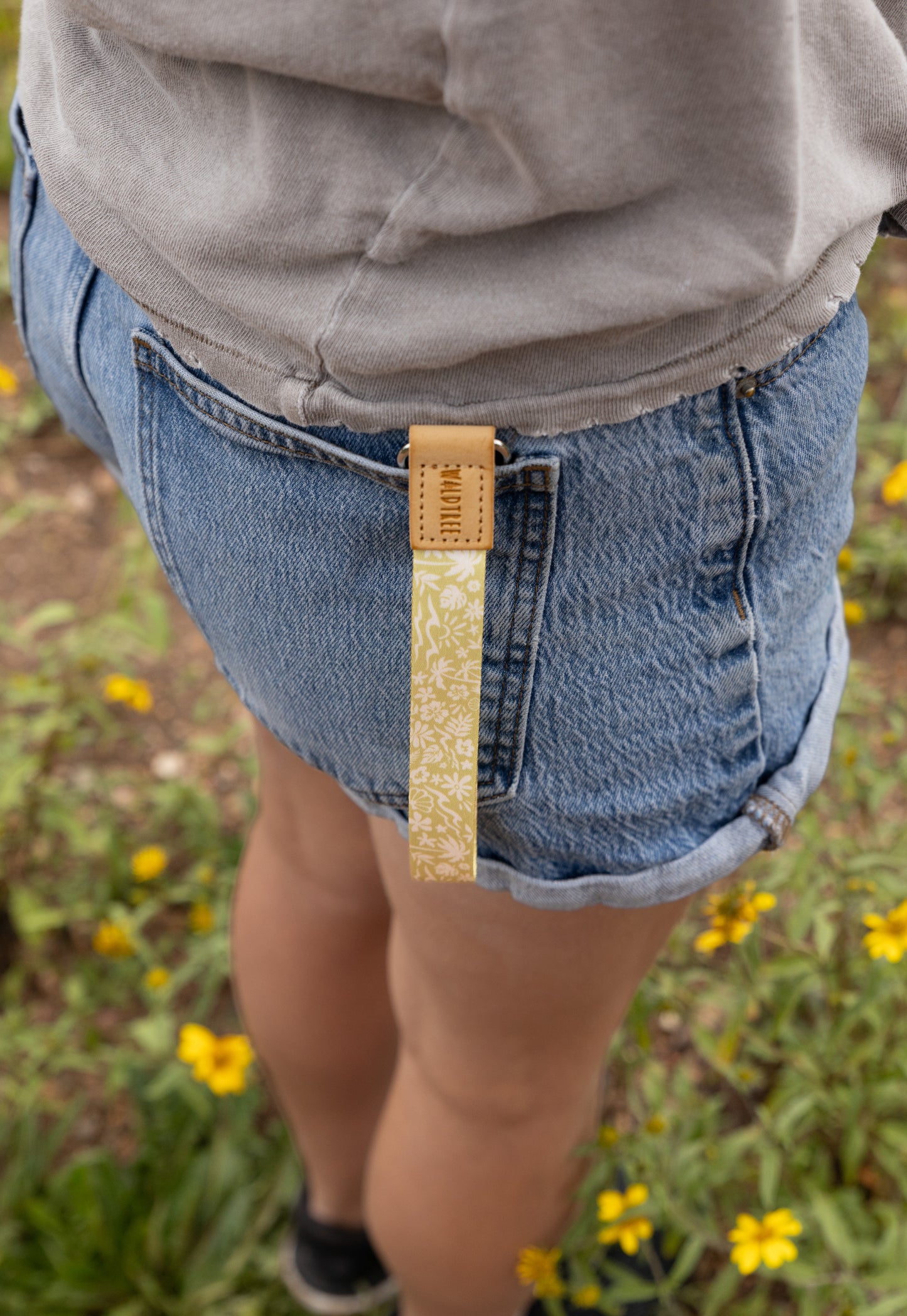 The Sunny Day wristlet keychain clipped to the back belt loop of denim shorts, the yellow strap printed with a beach-themed pattern of waves, palm trees, and seashells hanging against the wearer's leg in a field of yellow wildflowers.