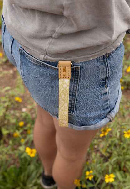 The Sunny Day wristlet keychain clipped to the back belt loop of denim shorts, the yellow strap printed with a beach-themed pattern of waves, palm trees, and seashells hanging against the wearer's leg in a field of yellow wildflowers.