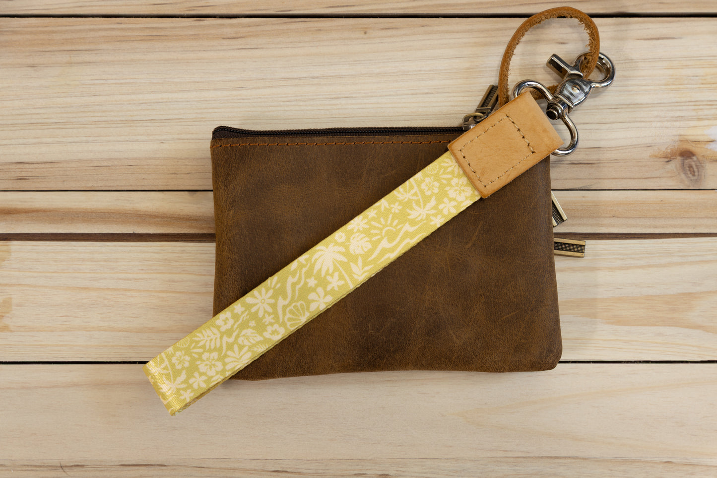 The Sunny Day wristlet keychain attached to a set of keys and draped across a brown leather zip pouch on a wood surface, showing the yellow strap printed with waves, palm trees, and seashells, and the tan leather WILDTREE tab.