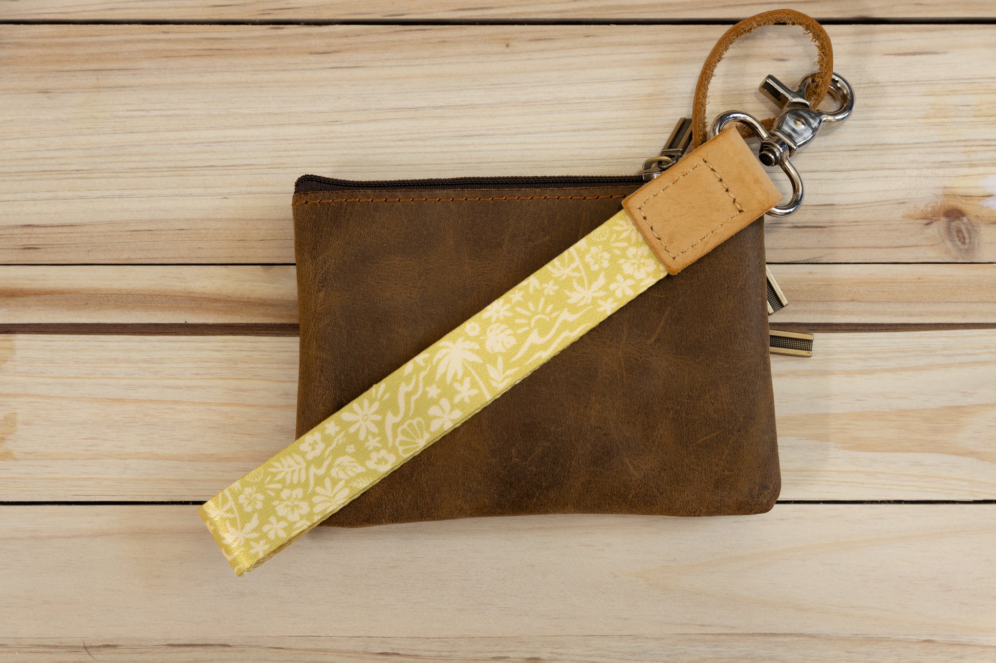 The Sunny Day wristlet keychain attached to a set of keys and draped across a brown leather zip pouch on a wood surface, showing the yellow strap printed with waves, palm trees, and seashells, and the tan leather WILDTREE tab.