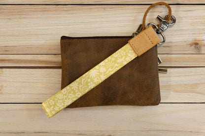 The Sunny Day wristlet keychain attached to a set of keys and draped across a brown leather zip pouch on a wood surface, showing the yellow strap printed with waves, palm trees, and seashells, and the tan leather WILDTREE tab.