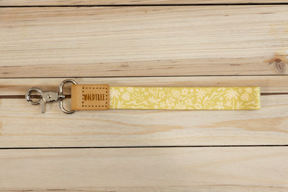 Close-up of the Sunny Day wristlet keychain laid flat on a wood plank surface, showing the yellow strap printed with a detailed white pattern of waves, palm trees, suns, flowers, and seashells, and the tan leather WILDTREE tab.