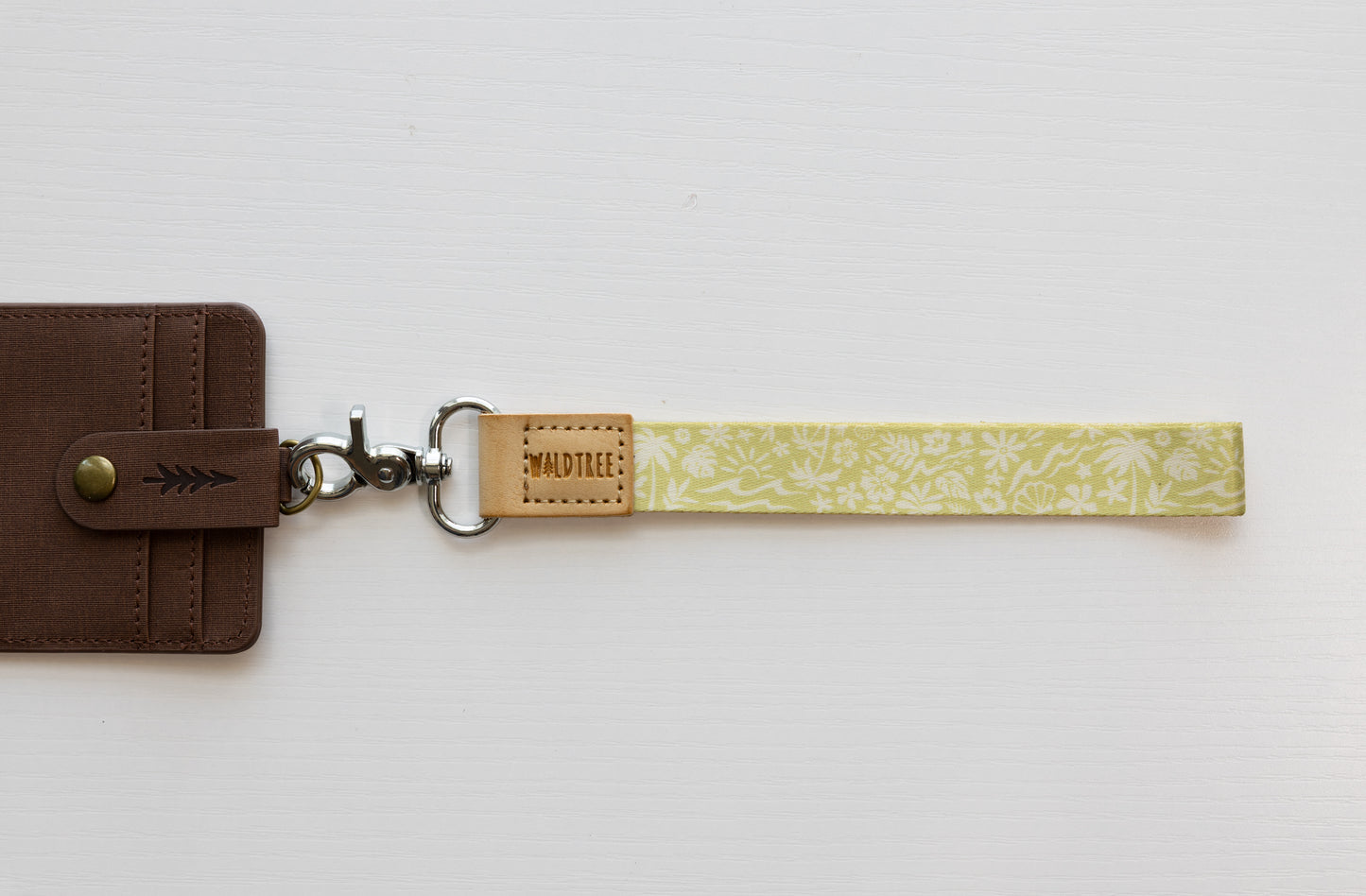 The Sunny Day wristlet keychain attached to a brown vegan-leather bifold wallet on a white background, showing the yellow strap printed with waves, palm trees, and seashells, the tan leather WILDTREE tab, and a silver swivel clasp.