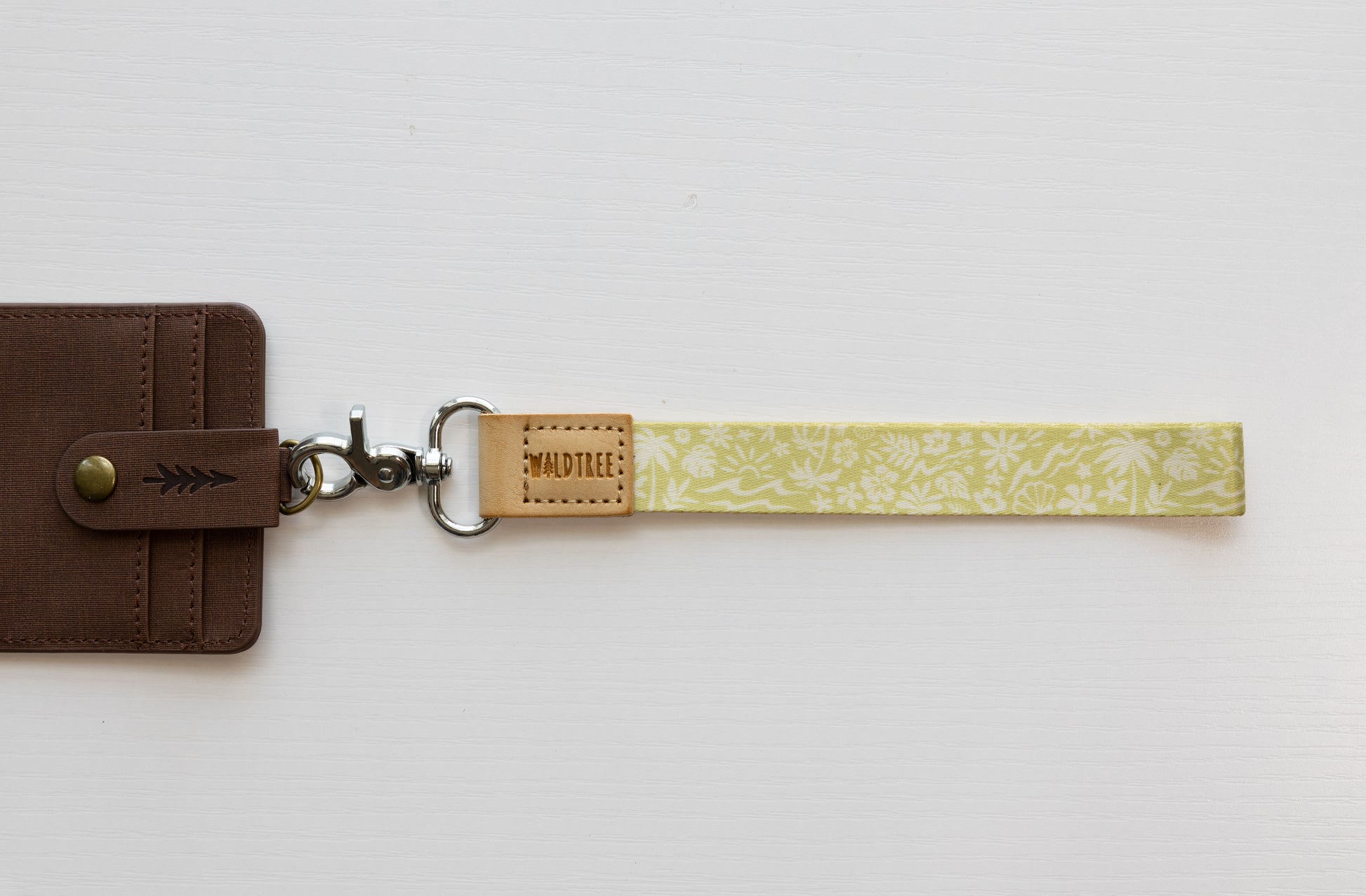 The Sunny Day wristlet keychain attached to a brown vegan-leather bifold wallet on a white background, showing the yellow strap printed with waves, palm trees, and seashells, the tan leather WILDTREE tab, and a silver swivel clasp.