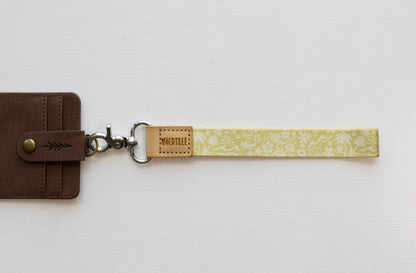 The Sunny Day wristlet keychain attached to a brown vegan-leather bifold wallet on a white background, showing the yellow strap printed with waves, palm trees, and seashells, the tan leather WILDTREE tab, and a silver swivel clasp.