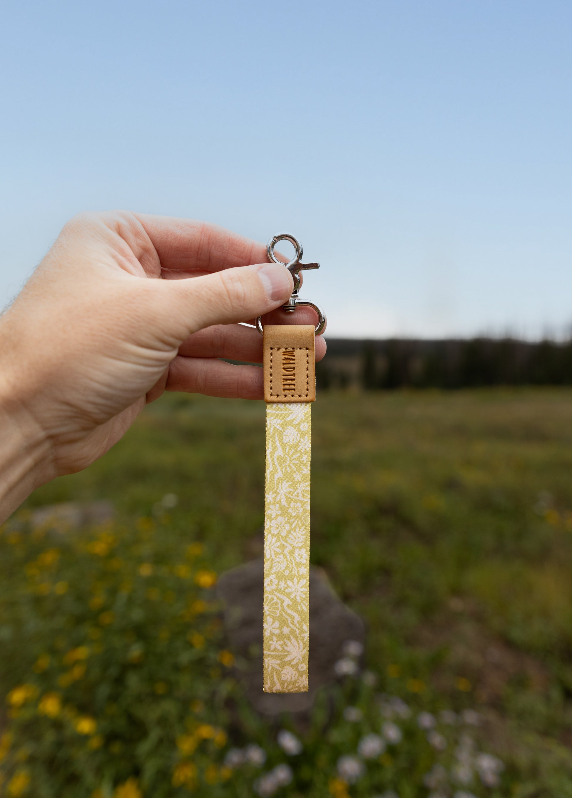 A hand holds the Sunny Day wristlet keychain up against a blue sky and grassy wildflower field, showing the yellow strap printed with waves, palm trees, and seashells, the tan leather WILDTREE tab, and silver clasps.