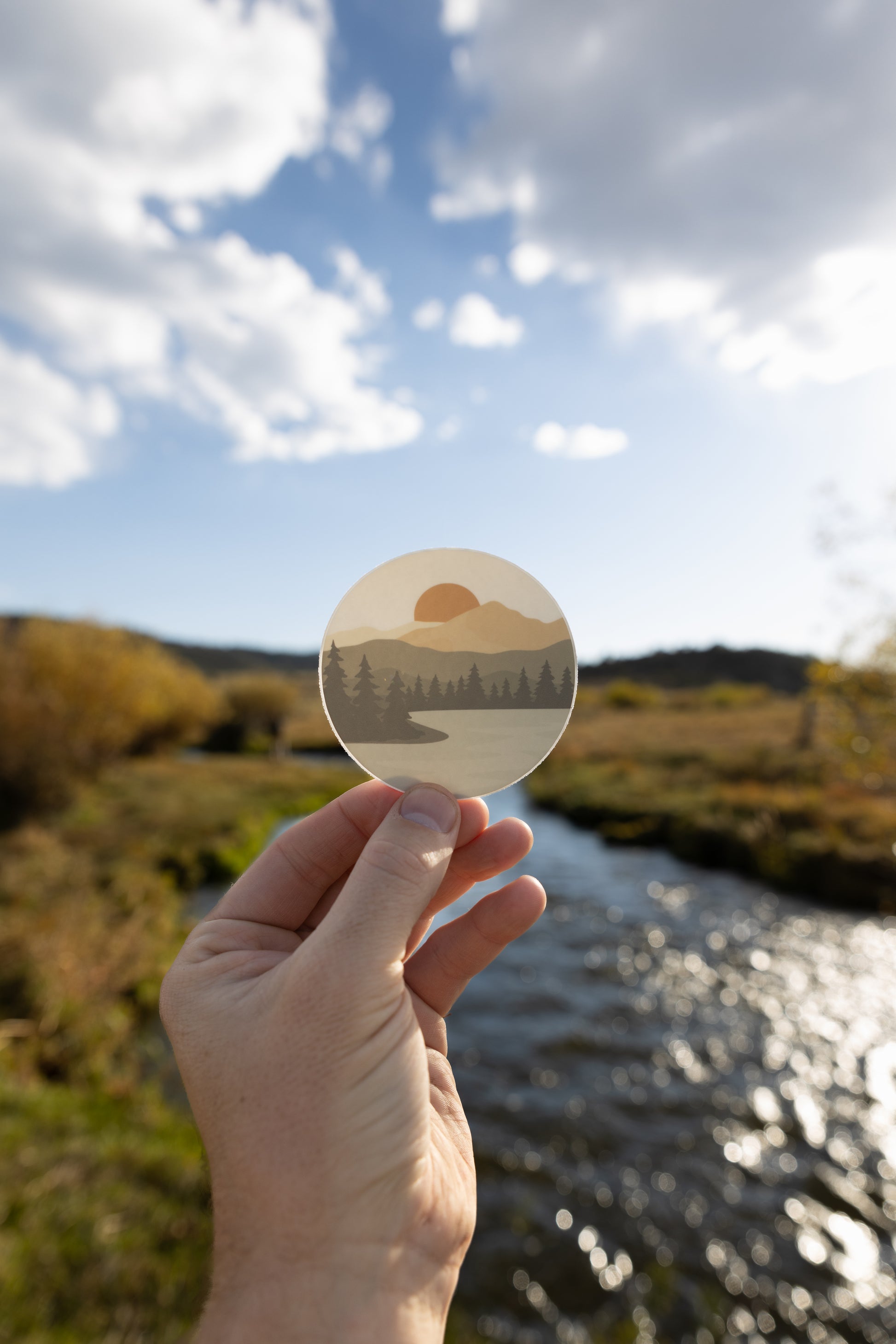 Hand holding up  Sunrise Lake Sticker featuring lake, mountains, trees and sun in front of river and valley