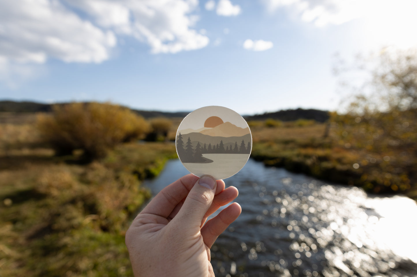 Hand holding up  Sunrise Lake Sticker featuring lake, mountains, trees and sun in front of river and valley