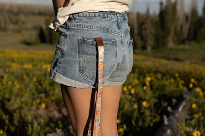 A person wearing denim shorts and a white top stands in a field of yellow wildflowers. The long Sunset Floral Neck Lanyard is clipped to a belt loop on the back of their shorts, hanging down the back of their leg.