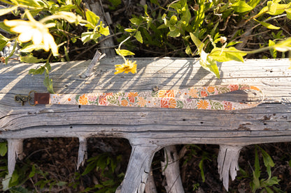 The Sunset Floral Neck Lanyard, featuring a pattern of orange, purple, and green flowers on a light background, is laid horizontally on a weathered gray log. The full length of the lanyard strap, the brown leather tag, and the brass clasp are visible. Yellow wildflowers and green foliage are in the sunny background.