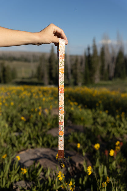 A person's hand holds the long Sunset Floral Neck Lanyard vertically against a blue sky and a blurred outdoor landscape of yellow wildflowers and evergreen trees. The lanyard has a colorful floral print, a brown leather end, and a brass clasp.