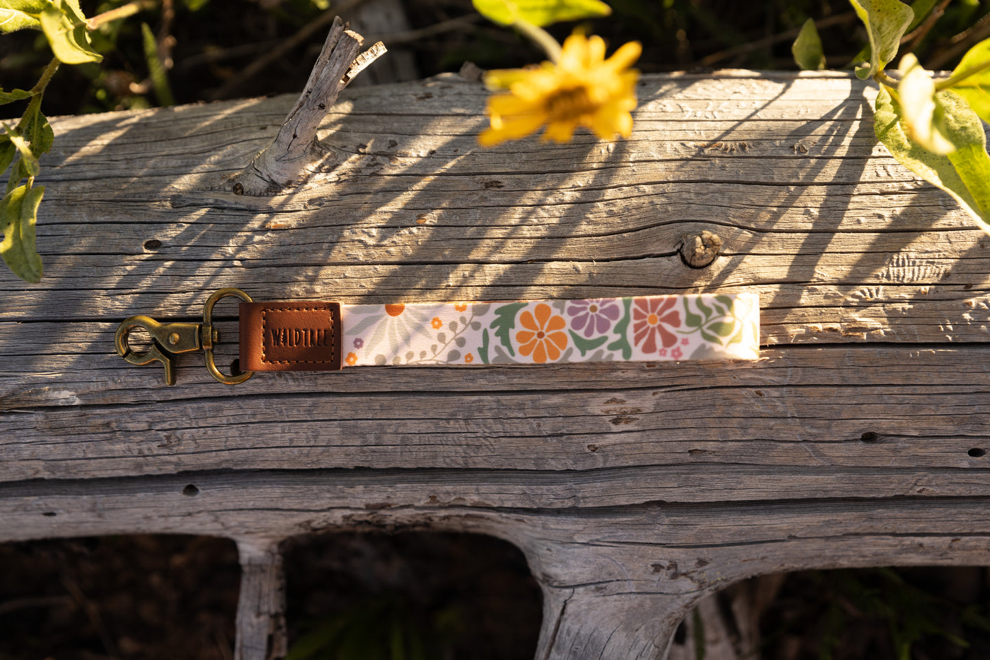 The Sunset Floral wristlet keychain, featuring a pattern of orange, purple, and green flowers on a light background, is laid horizontally on a weathered gray log. The keychain has a brown leather tag stamped with "WILDTREE" and a brass lobster claw clasp. A blurred yellow wildflower hangs down from the top.