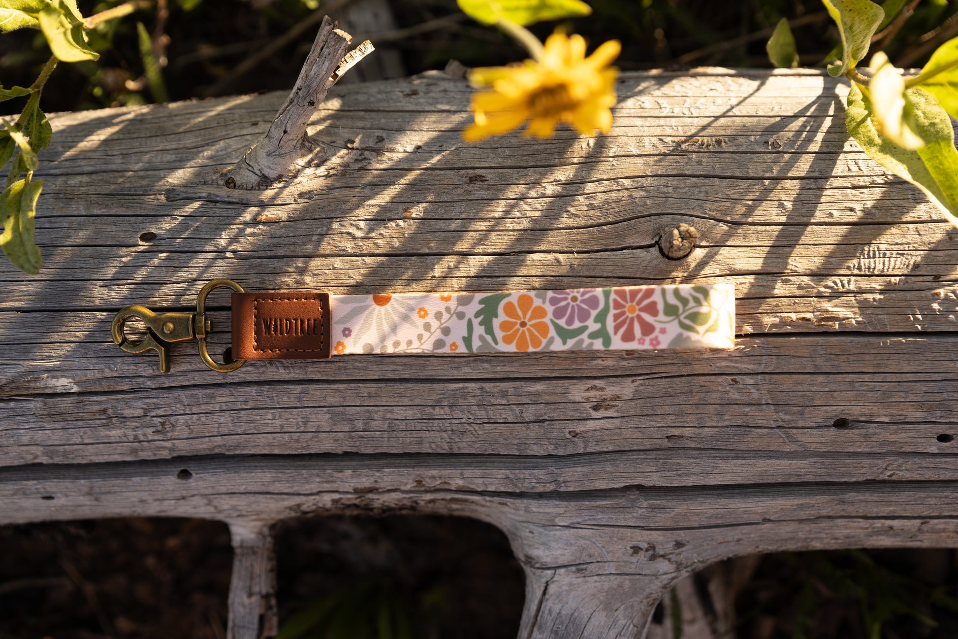 The Sunset Floral wristlet keychain, featuring a pattern of orange, purple, and green flowers on a light background, is laid horizontally on a weathered gray log. The keychain has a brown leather tag stamped with "WILDTREE" and a brass lobster claw clasp. A blurred yellow wildflower hangs down from the top.