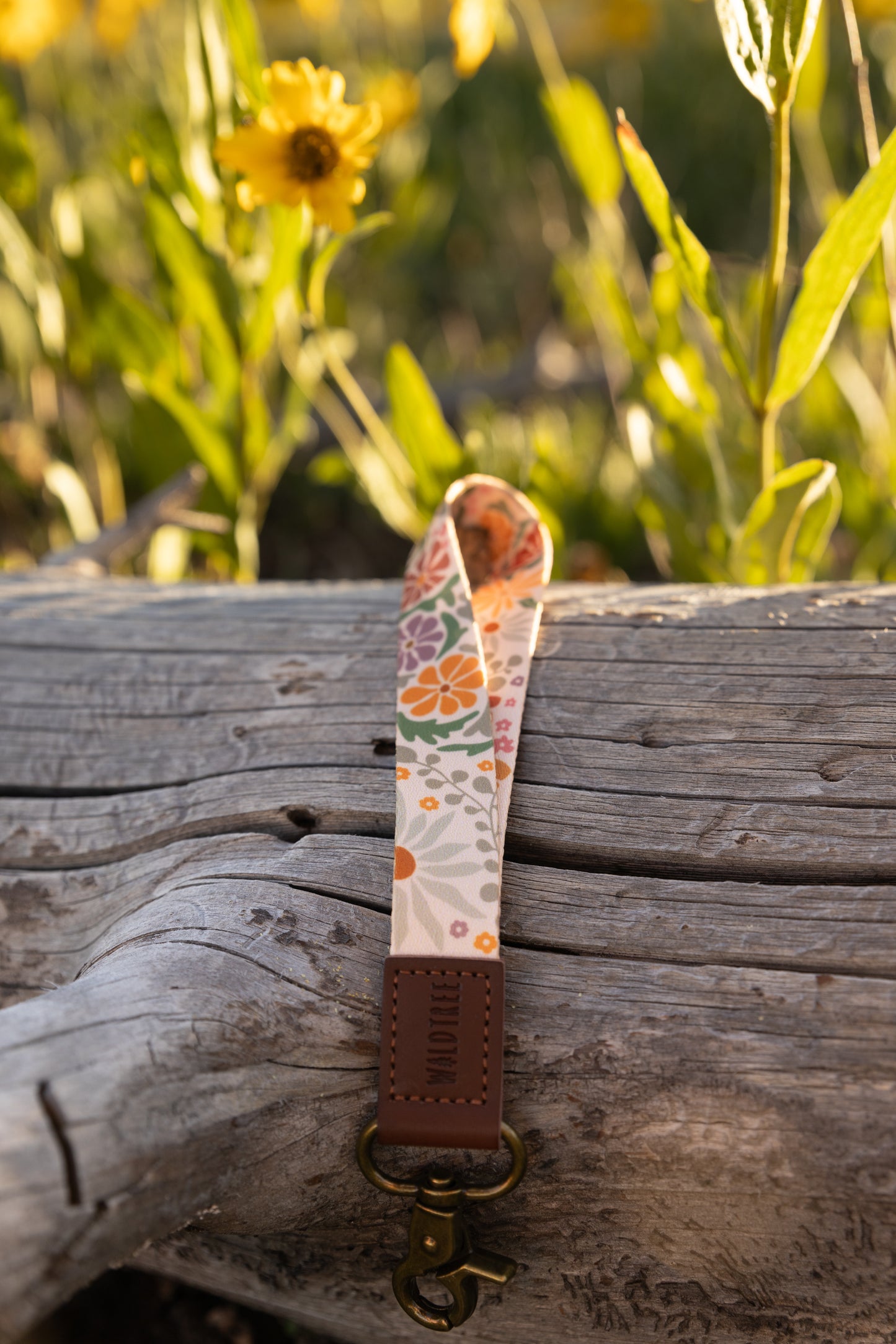 The Sunset Floral wristlet keychain is positioned vertically on a textured gray log. The colorful floral strap is looped at the top, and the bottom shows the brown leather tag and brass clasp. Bright green foliage and a yellow flower are in the blurry background.