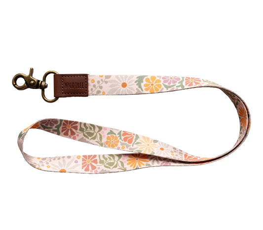 "Wildtree sunset flower neck lanyard" on white. Light tan strap with orange, pink, and muted green stylized flowers. Brown leather loop ("WILDTREE") attaches to a bronze swivel clasp. Curved to display the vibrant floral design.