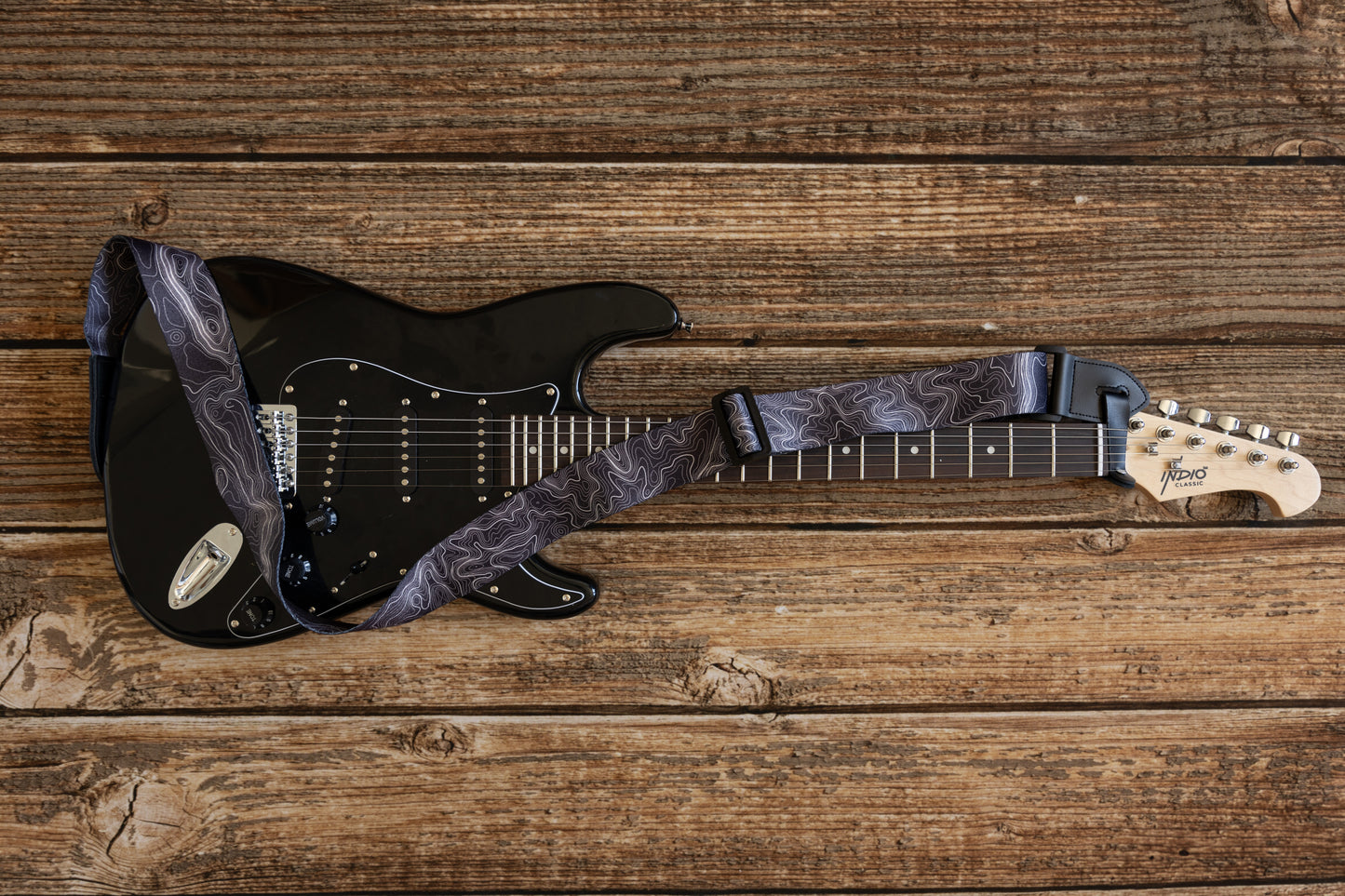 Topographic map designed black and white guitar strap connected to black electric guitar laying on wood floor