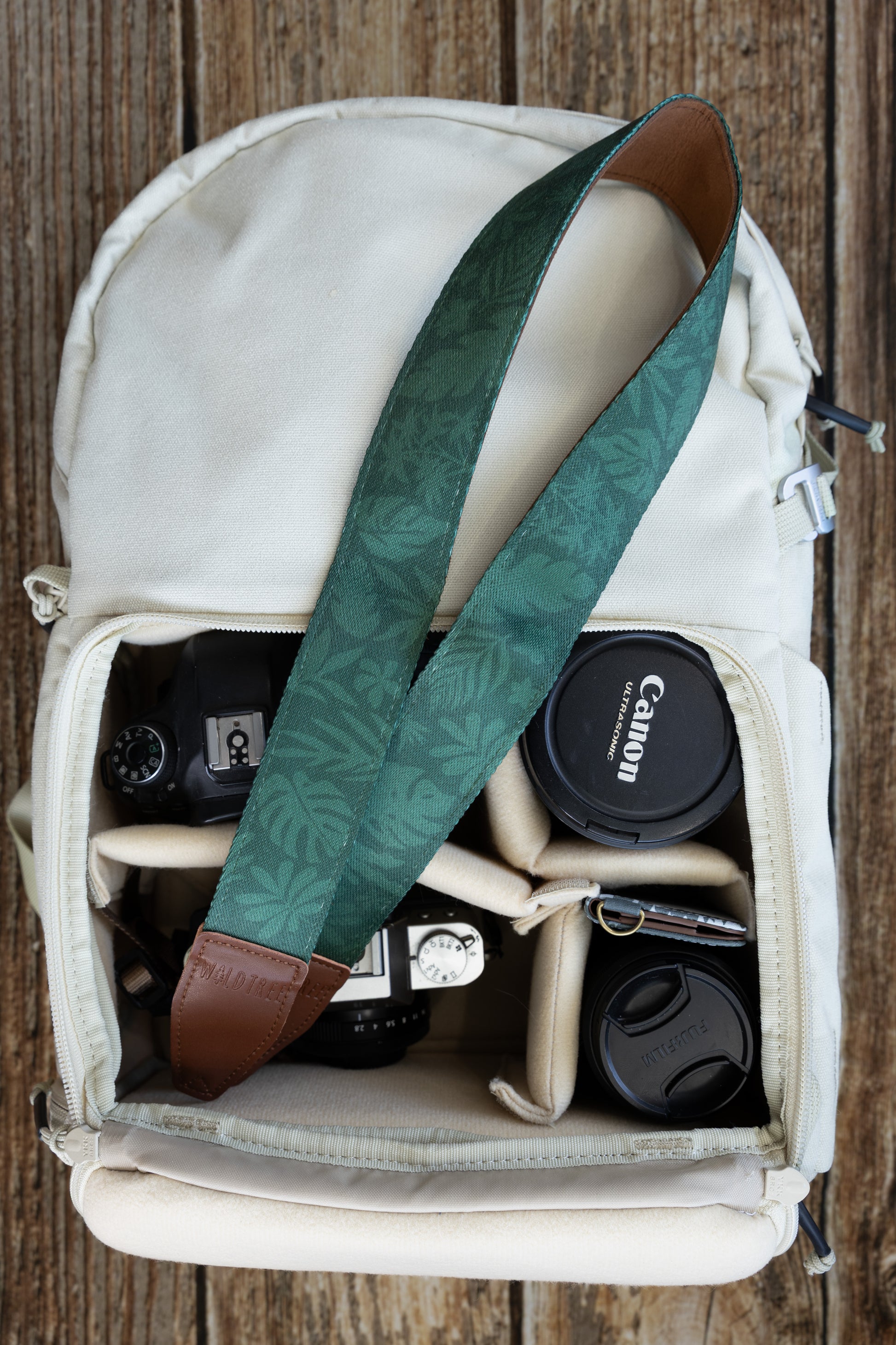 op-down view inside a cream-colored camera bag showing a WILDTREE camera strap resting across the top, featuring a repeating tropical leaf pattern in shades of green, placed above a camera body and lenses.
