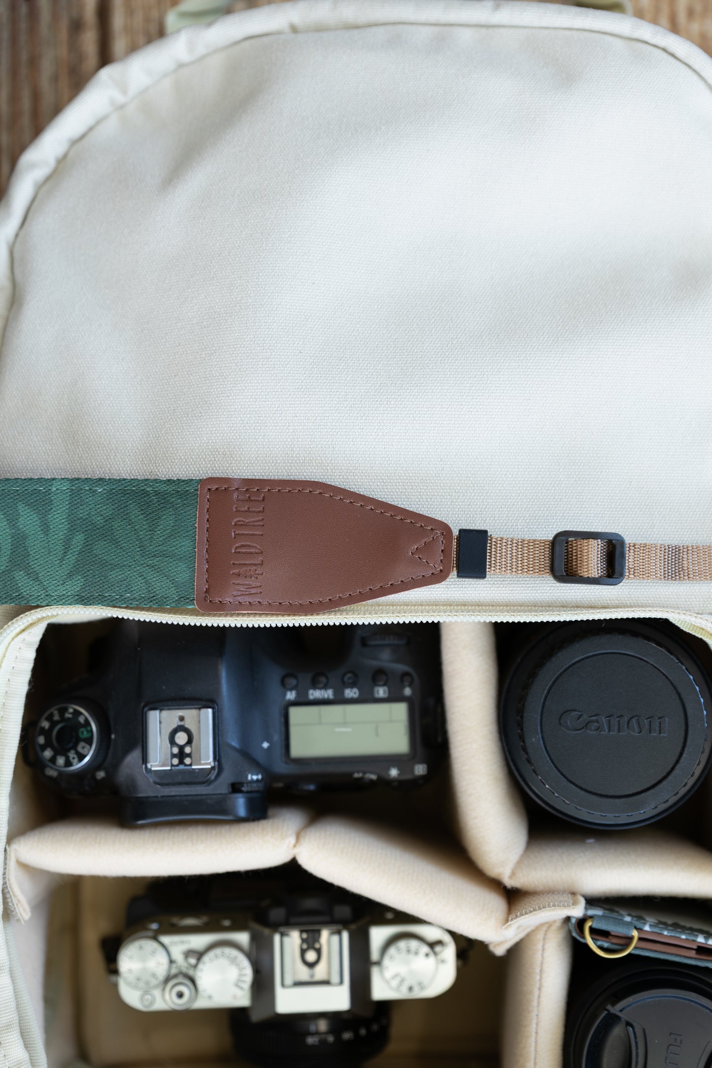Top-down view inside a cream-colored camera bag showing a WILDTREE camera strap resting across the top, featuring a dark green botanical leaf print and brown vegan leather attachment ends, positioned above a camera body and lenses.