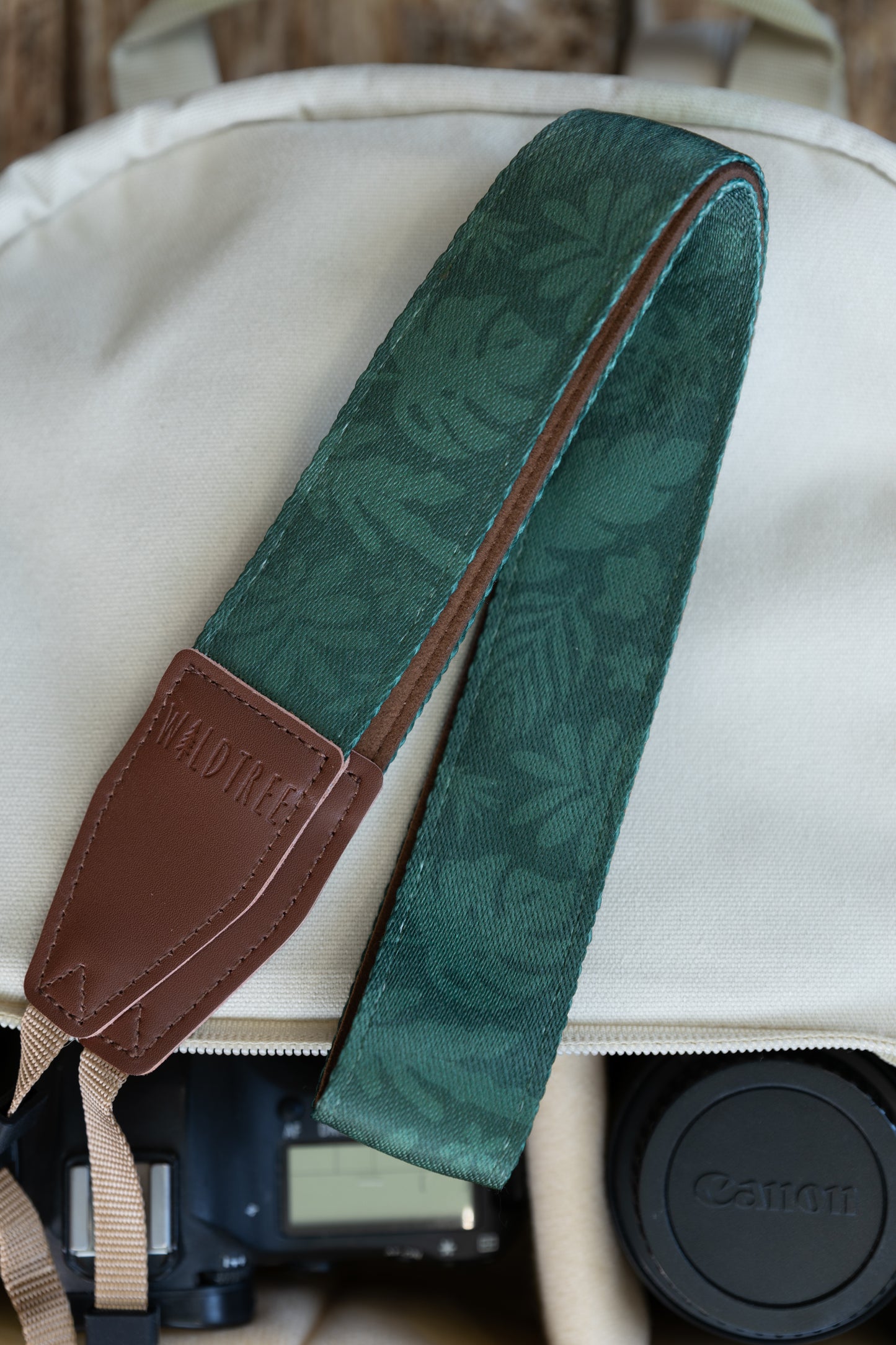 Top-down view of the WILDTREE camera strap coiled inside a cream-colored camera bag, highlighting the contrast between the green tropical leaf pattern and the branded brown vegan leather attachment end.