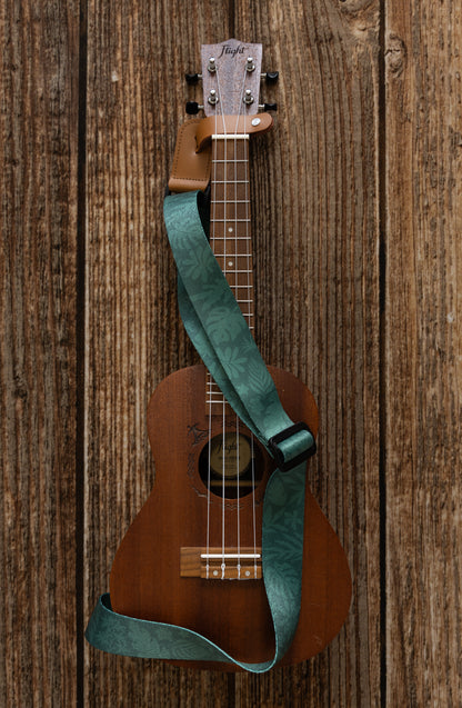 Green tropical leaf printed Ukulele Strap attached to Ukulele lying on wood floor