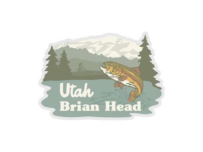 This sticker design for Brian Head, Utah features an outdoor scene with a rainbow trout jumping out of the water in the foreground. The background includes snow-capped mountains, pine trees, and a small silhouette of a person on a bicycle near the trees. The text "Utah Brian Head" is overlaying the water.