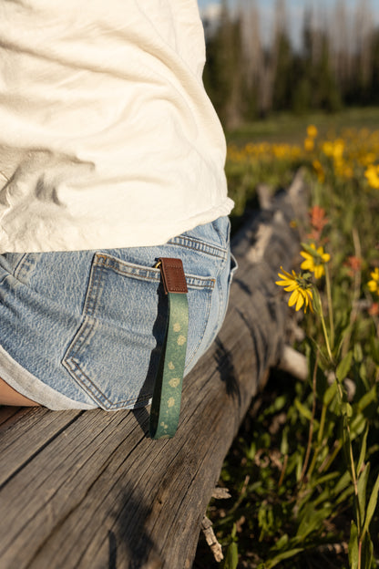 A green floral wristlet keychain hangs from the back pocket of a person’s denim shorts. The person is sitting on a weathered log in a field of yellow and orange wildflowers during the golden hour.
