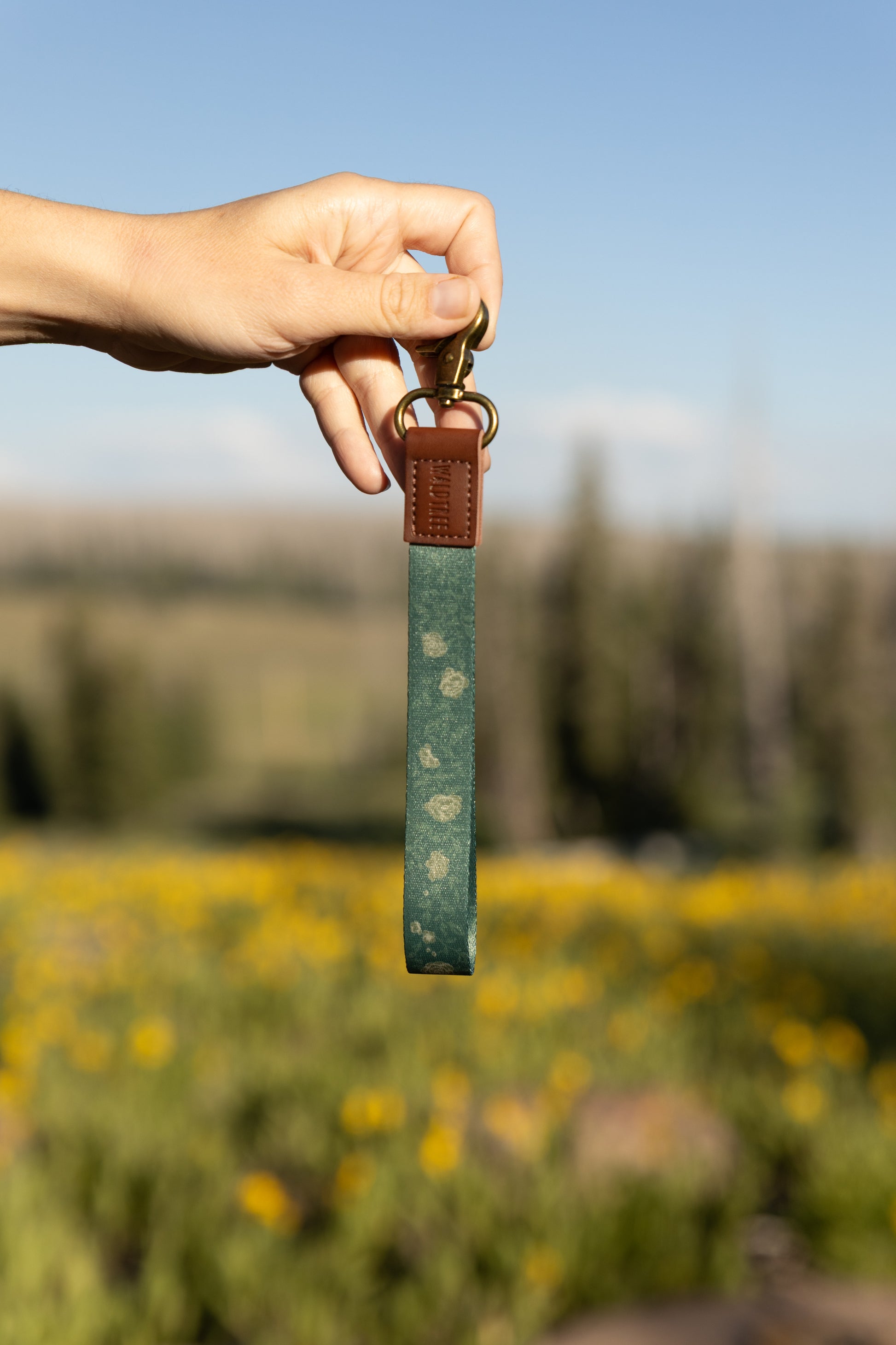 A hand holds a green floral wristlet keychain against a blurred background of a sunny field of yellow wildflowers and a blue sky. The wristlet features a muted rose pattern, brown leather trim, and a brass swivel snap hook.