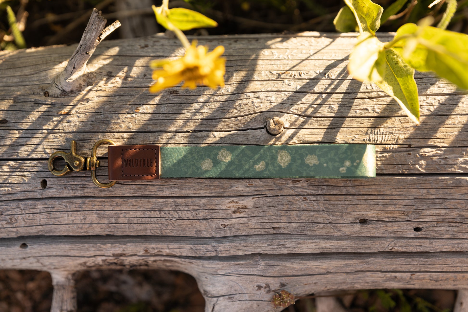 A top-down view of a green floral wristlet keychain resting on a textured, sunlit fallen log. The keychain features a pattern of small white roses, a brown leather accent with the "WILDTREE" logo, and a vintage-style brass clasp.