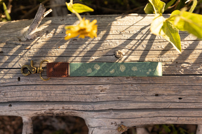A top-down view of a green floral wristlet keychain resting on a textured, sunlit fallen log. The keychain features a pattern of small white roses, a brown leather accent with the "WILDTREE" logo, and a vintage-style brass clasp.