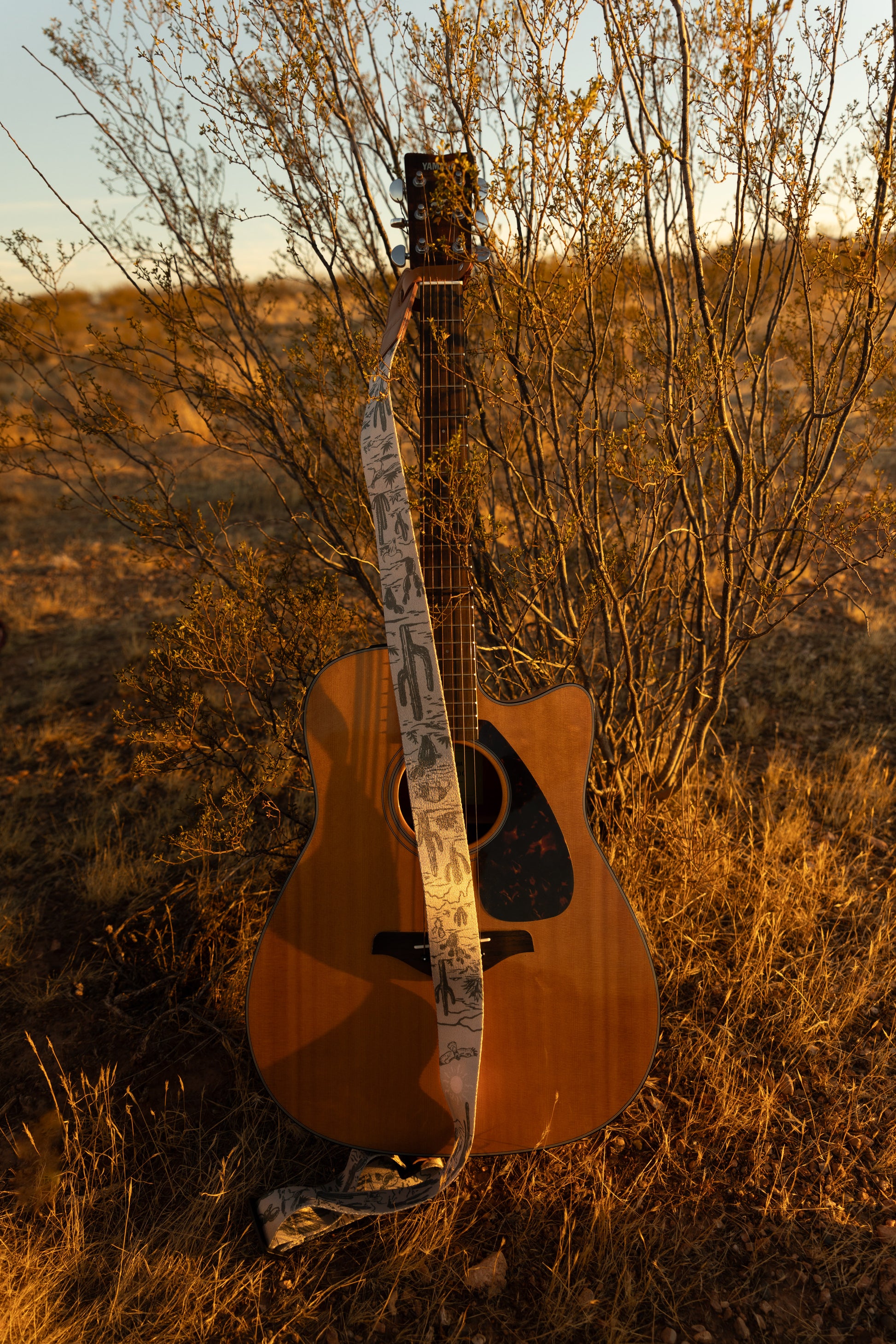 Western landscape guitar strap connected to acoustic guitar lying against desert bush. Tan strap printed with green desert mountains, wildlife, cacti, bones, birds, snakes, plants