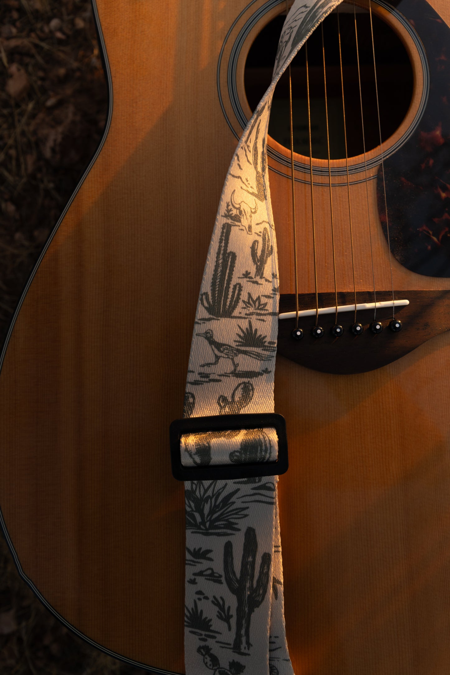 close up of Western landscape guitar strap connected to acoustic guitar lying on desert ground. Tan strap printed with green desert mountains, wildlife, cacti, bones, birds, snakes, plants