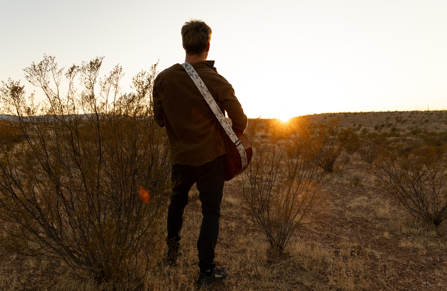 man standing in desert playing guitar wearing Western landscape guitar strap. Tan strap printed with green desert mountains, wildlife, cacti, bones, birds, snakes, plants