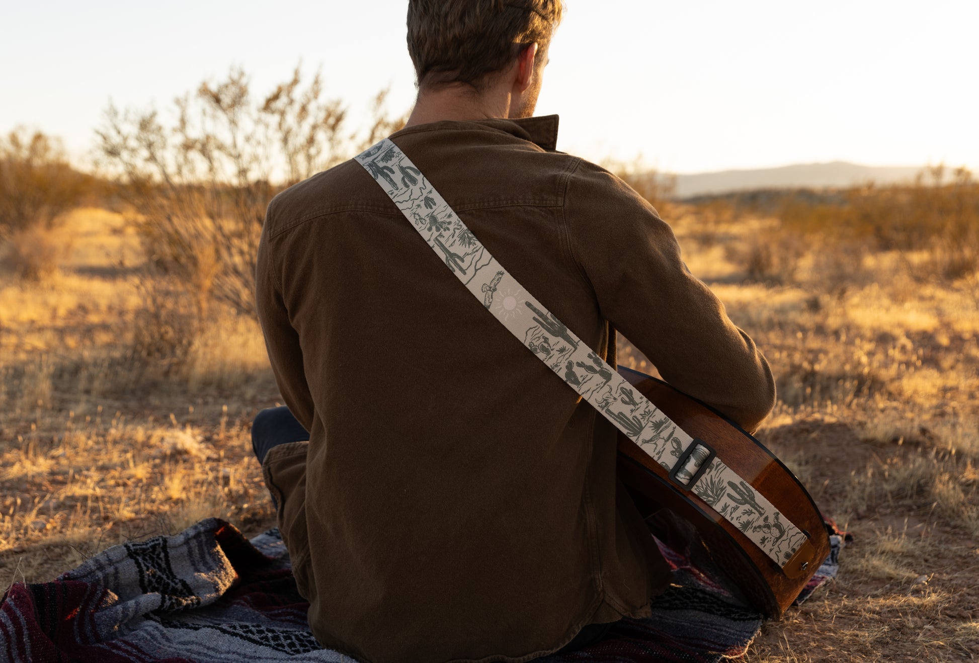 man sitting on desert ground playing guitar wearing Western landscape guitar strap. Tan strap printed with green desert mountains, wildlife, cacti, bones, birds, snakes, plants