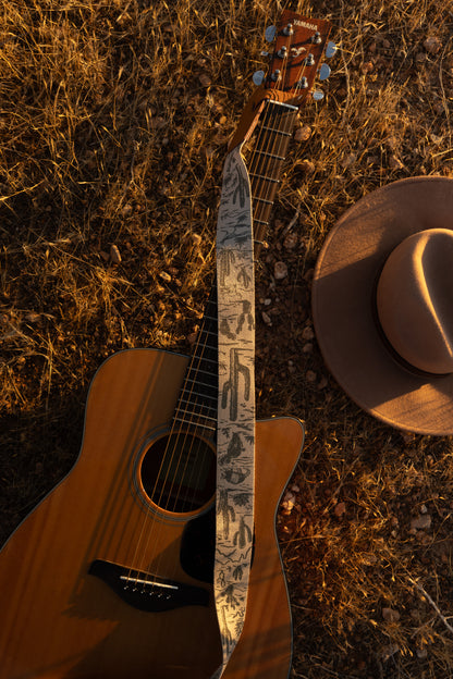 Western landscape guitar strap connected to acoustic guitar lying on desert ground. Tan strap printed with green desert mountains, wildlife, cacti, bones, birds, snakes, plants