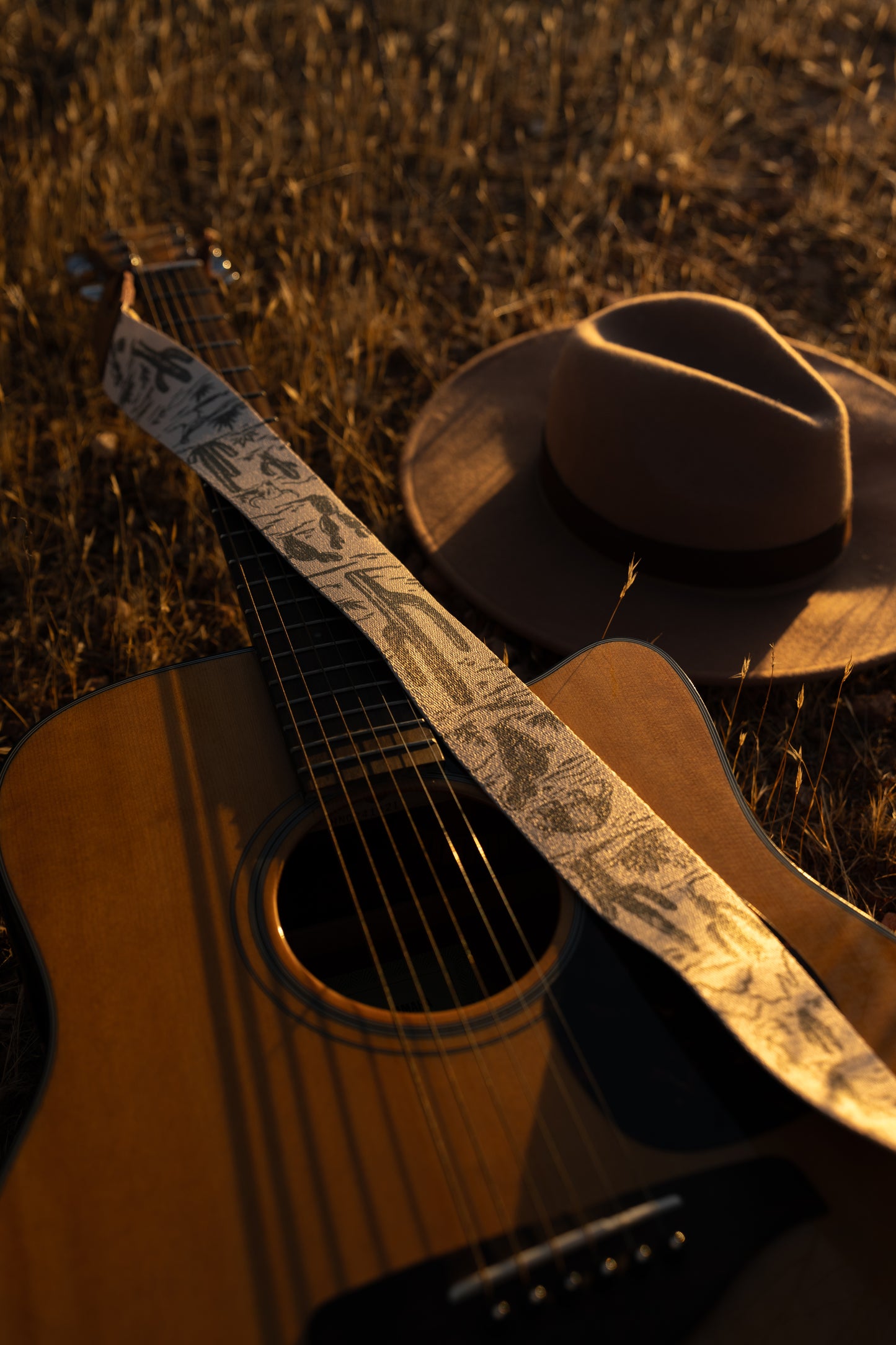 Western landscape guitar strap connected to acoustic guitar lying on desert ground. Tan strap printed with green desert mountains, wildlife, cacti, bones, birds, snakes, plants