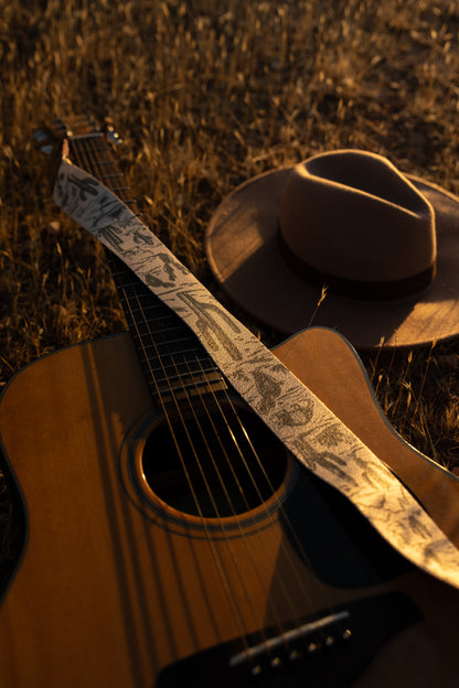 Western landscape guitar strap connected to acoustic guitar lying on desert ground. Tan strap printed with green desert mountains, wildlife, cacti, bones, birds, snakes, plants