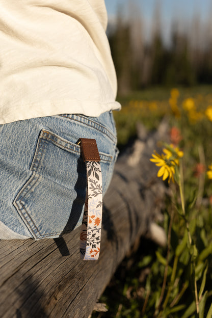 A person sits on a weathered log, and a wristlet keychain with a dark floral and foliage pattern hangs from a belt loop on their denim shorts. A bright yellow wildflower is visible in the foreground to the right.