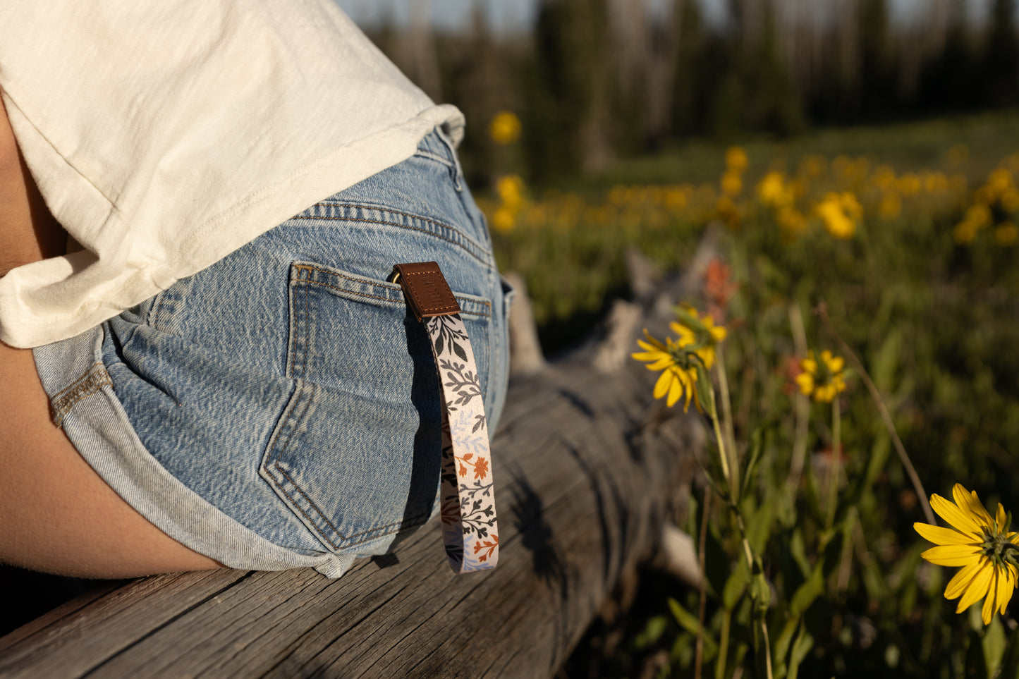 A person sitting on a weathered log outdoors, wearing denim shorts and a white top. A wristlet keychain with a light-colored background and a pattern of dark green, rust, and light blue foliage hangs from a belt loop on the back pocket. Yellow wildflowers are in the blurry foreground and background.