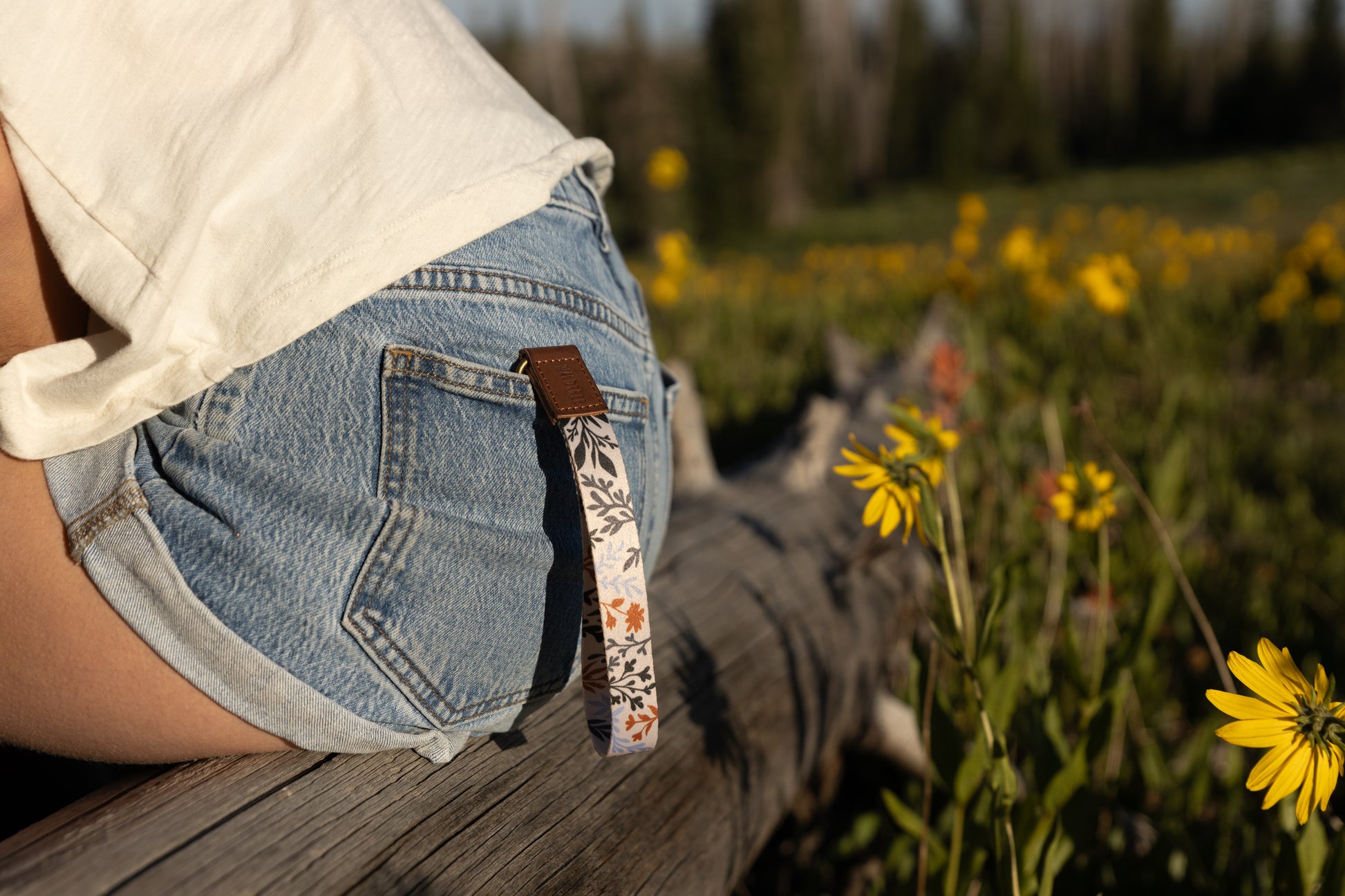 A person sitting on a weathered log outdoors, wearing denim shorts and a white top. A wristlet keychain with a light-colored background and a pattern of dark green, rust, and light blue foliage hangs from a belt loop on the back pocket. Yellow wildflowers are in the blurry foreground and background.