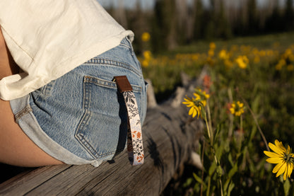 A person sitting on a weathered log outdoors, wearing denim shorts and a white top. A wristlet keychain with a light-colored background and a pattern of dark green, rust, and light blue foliage hangs from a belt loop on the back pocket. Yellow wildflowers are in the blurry foreground and background.