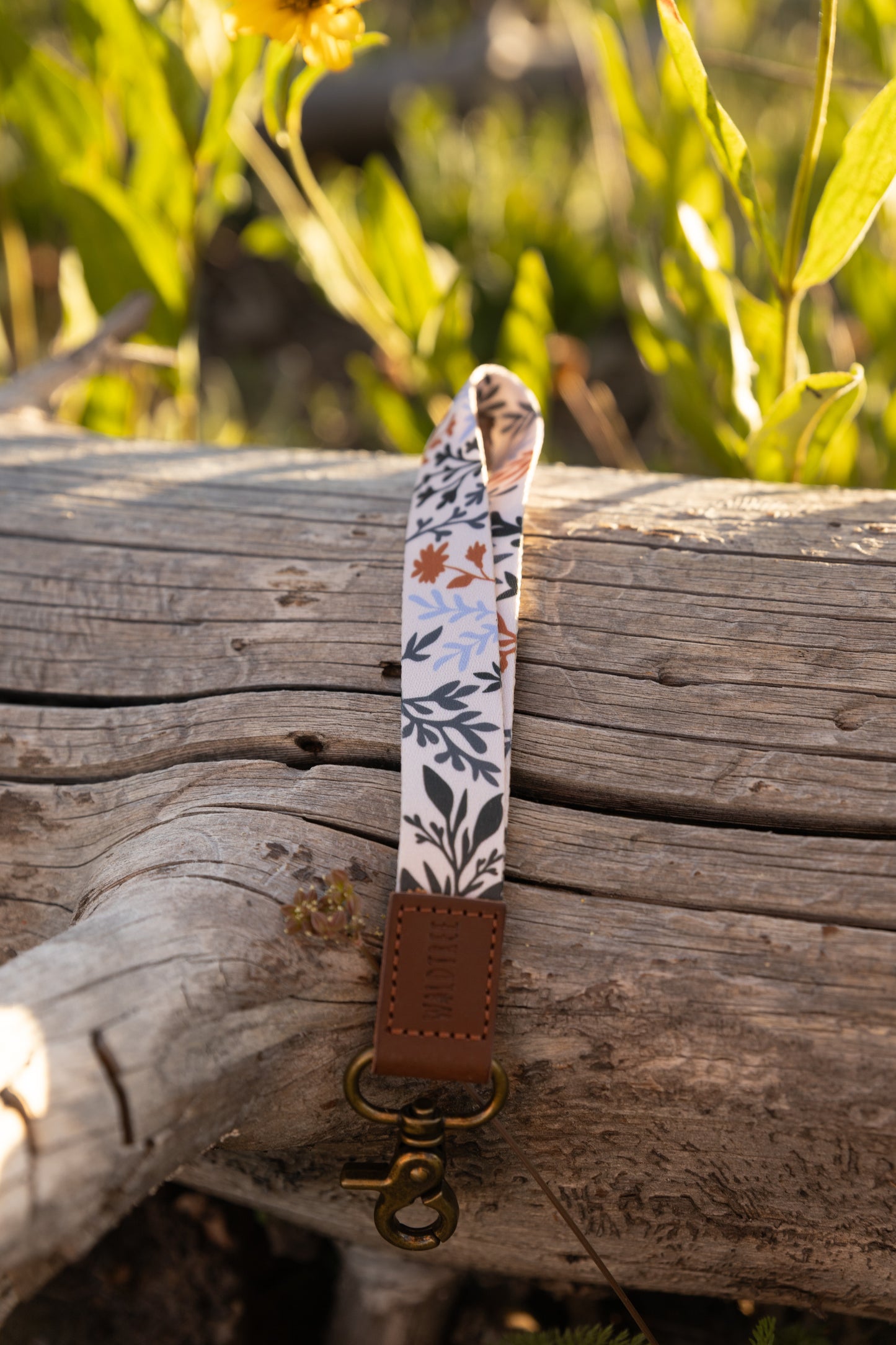 A close-up, vertical shot of the wildflower-patterned wristlet keychain resting on a gray, textured log. The pattern features dark green, brown, and light blue leaves and stems. The keychain has a brown leather tag and a brass clasp. Bright foliage is in the blurred background.