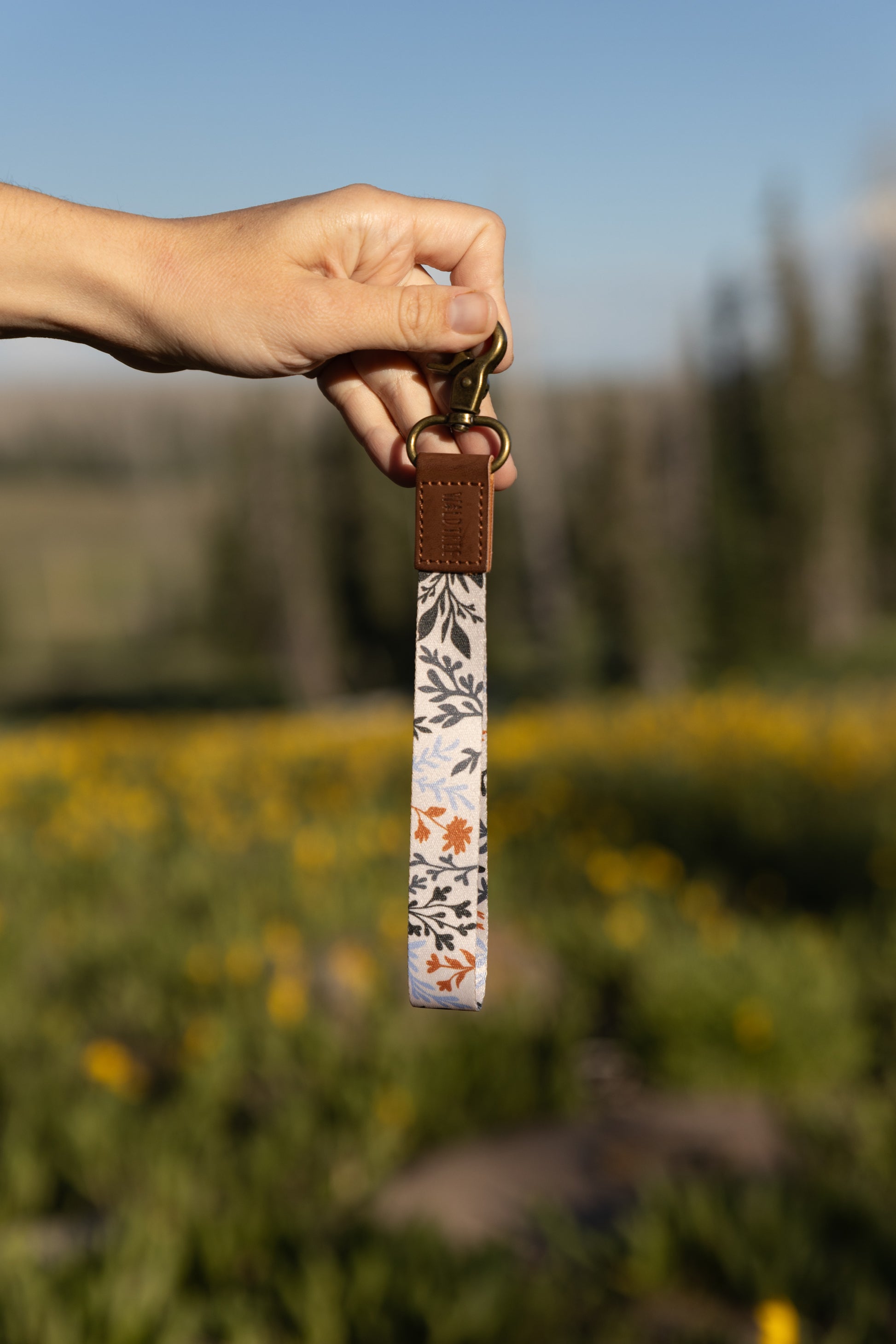 A hand holds up a wristlet keychain featuring a pattern of dark green, rust, and light blue foliage on a light background. The keychain has a brown leather end and a brass clasp. The background is a bright, sunny, out-of-focus field of yellow flowers and green foliage.