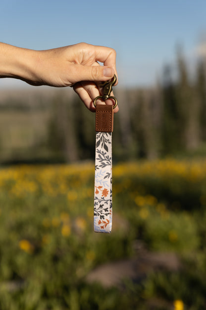 A hand holds up a wristlet keychain featuring a pattern of dark green, rust, and light blue foliage on a light background. The keychain has a brown leather end and a brass clasp. The background is a bright, sunny, out-of-focus field of yellow flowers and green foliage.