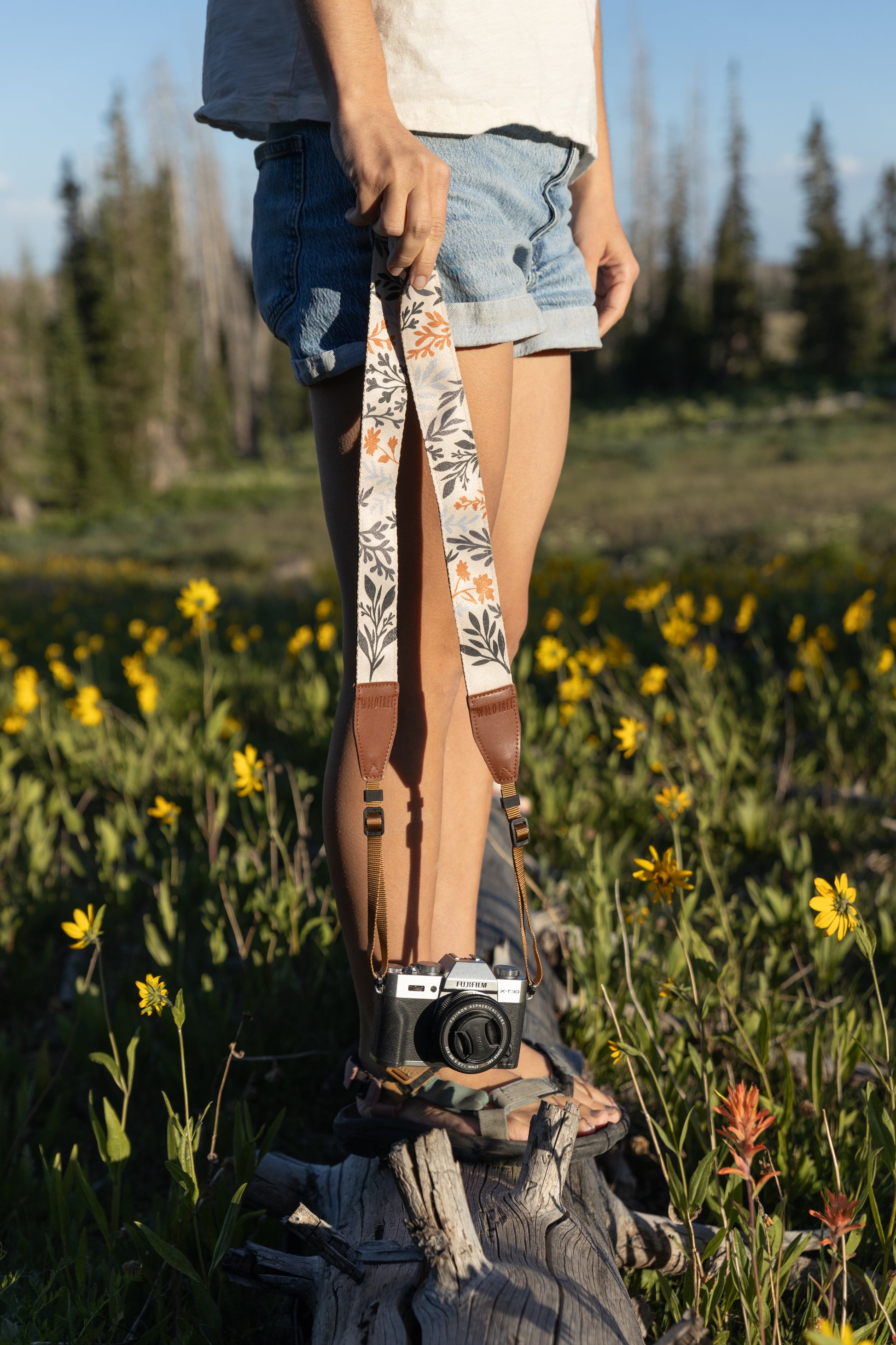 A person in a white shirt and denim shorts stands on a weathered log in a field of yellow and orange wildflowers. They hold a Wildtree Outdoor camera strap featuring a white and cream fabric with a detailed wildflower print in deep greens, grays, and rust orange, connected to a brown leather strap and a black and silver camera.