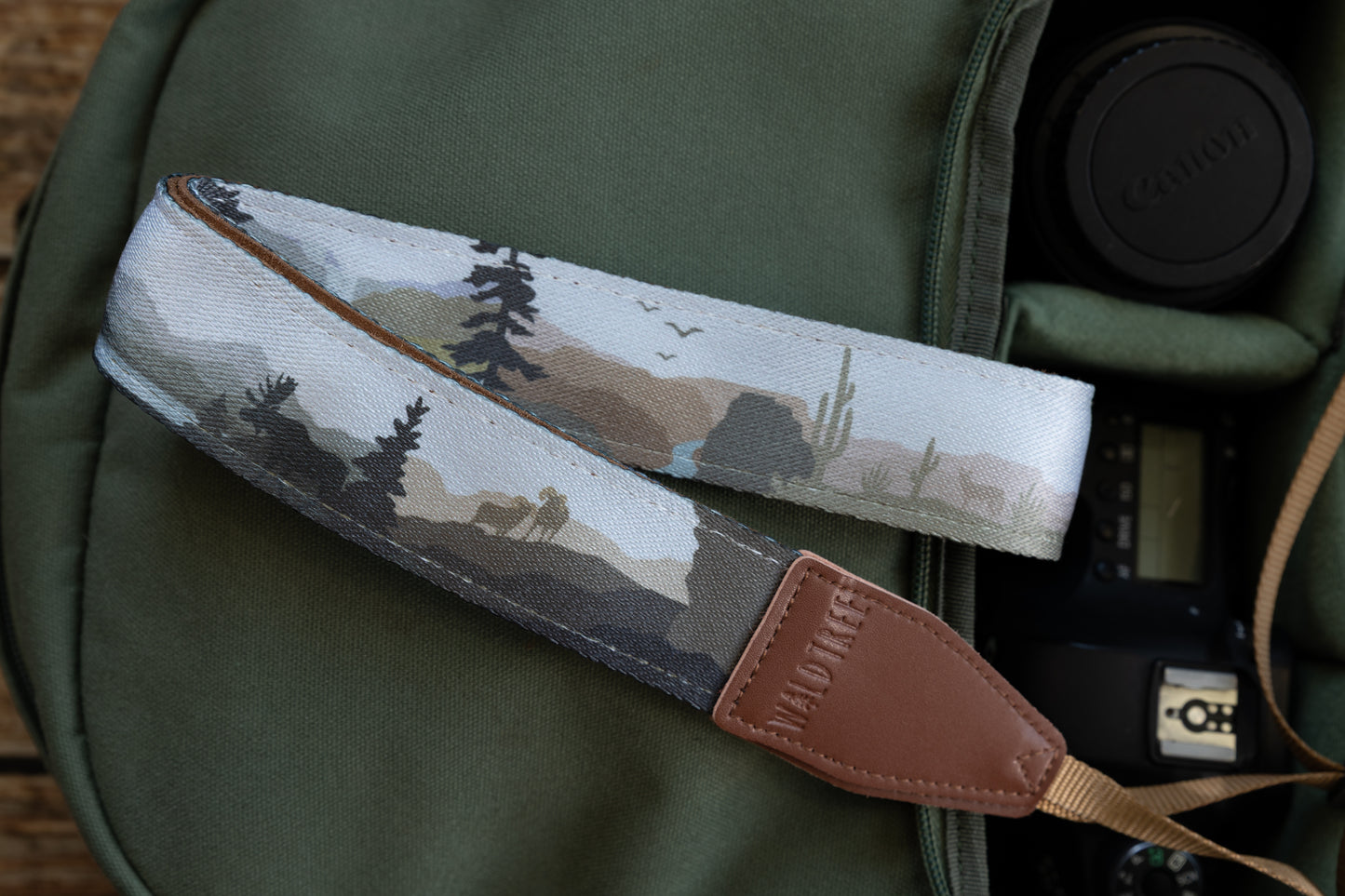 A folded camera strap displaying a collage of nature scenes, including a bear, desert plants, and mountain ranges, with brown leather attachments branded "WILDTREE".