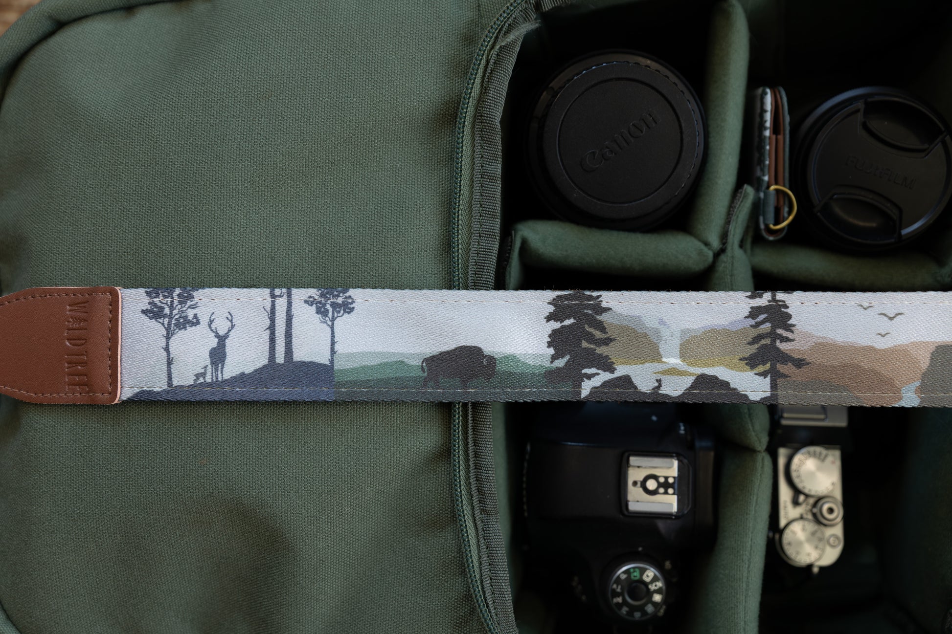 Close-up of a camera strap highlighting a printed scene featuring a deer and fawn, a buffalo, and a distant waterfall set against a mountain and forest backdrop.