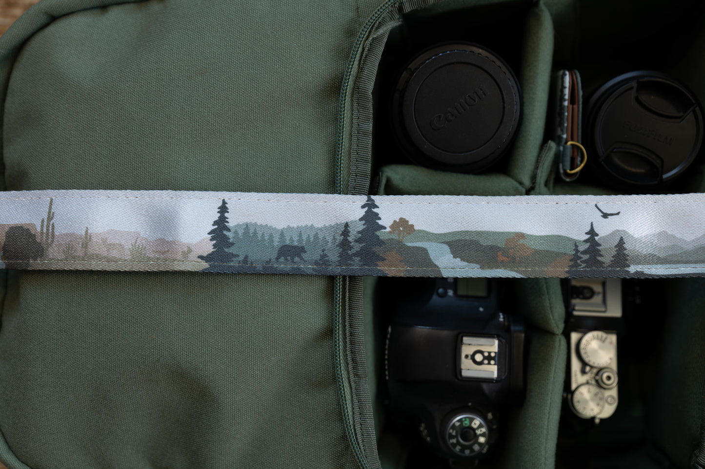 Close-up of a camera strap showing a nature design that transitions from a desert scene with a cactus to a forest with a bear silhouette, pine trees, and a flying eagle.