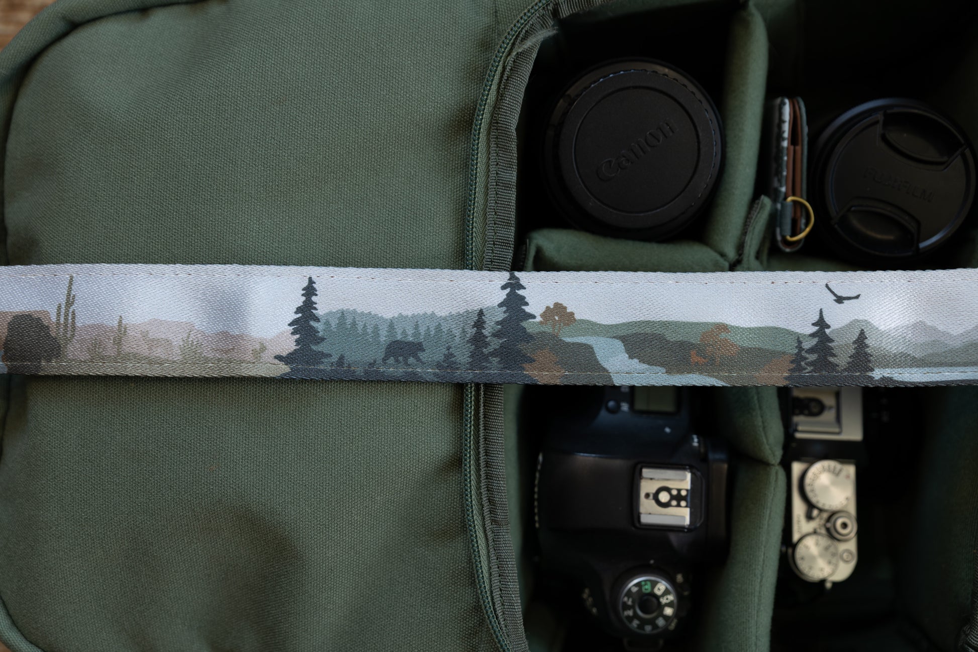 Close-up of a camera strap showing a nature design that transitions from a desert scene with a cactus to a forest with a bear silhouette, pine trees, and a flying eagle.