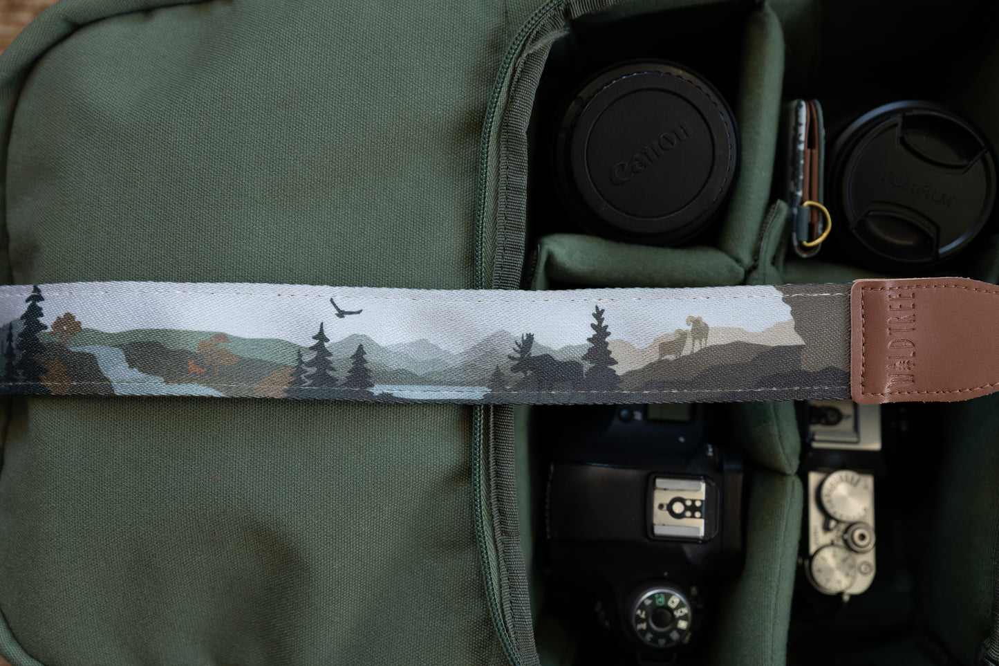 Close-up of a camera strap featuring a stylized nature illustration with pine trees, a moose silhouette, and an eagle flying over a mountain landscape, attached to a brown leather end piece with the logo "WILDTREE"